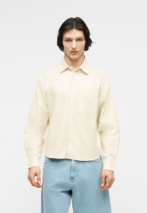 Νέος άνδρας με μεσαίου μήκους σκούρα μαλλιά, που φοράει κρεμ πουκάμισο με κουμπιά και ανοιχτόχρωμο τζιν, στέκεται μπροστά σε ένα απλό λευκό φόντο.