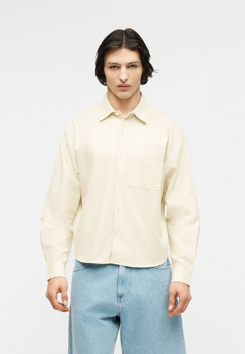 Νέος άνδρας με μεσαίου μήκους σκούρα μαλλιά, που φοράει κρεμ πουκάμισο με κουμπιά και ανοιχτόχρωμο τζιν, στέκεται μπροστά σε ένα απλό λευκό φόντο.