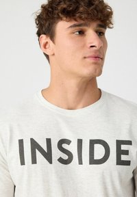 Camiseta de manga corta gris claro con texto negro en negrita que dice "INSIDE". Fabricada con una tela suave, con escote redondo y un corte holgado.