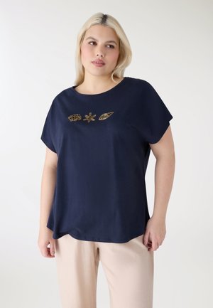 Donna che indossa una maglietta a maniche corte blu navy con ricami dorati di conchiglie, stelle marine e gusci, abbinata a pantaloni beige chiaro.