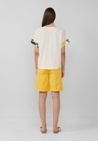 Witte t-shirt met gemusterde mouwen, gecombineerd met gele shorts. Het shirt heeft een losse pasvorm en omgeslagen zoom op de shorts. Eenvoudige sandalen maken de look compleet.