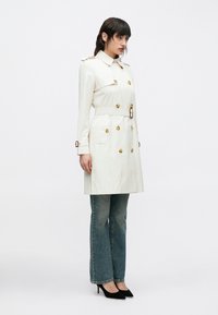 Krém színű ballonkabát arany gombokkal és övvel, bővülő szárú farmernadrággal és fekete, hegyes orrú magassarkú cipővel párosítva. Sima anyag, szabott fazon.