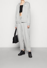 Šedá mikina na zip s malým logem, doplněná o odpovídající tepláky. Pod tím černý krátký top a černé tenisky. Doplněk: černá kabelka přes rameno.