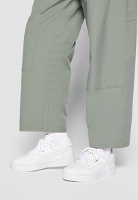 Baskets blanches avec texture perforée et accents dorés, associées à un pantalon vert clair à coupe ample avec des coutures visibles et des poches latérales.