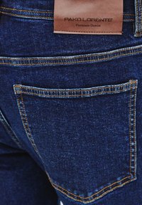 Ciemnoniebieskie dżinsy z kontrastującymi żółtymi szwami, z skórzaną metką oznaczoną "PAKO LORENTE" oraz detalem tylnej kieszeni.