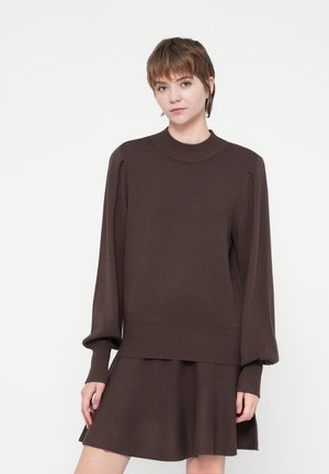 Jeune personne avec les cheveux bruns courts portant un pull brun à manches longues et une jupe brun assortie, sur fond blanc uni.