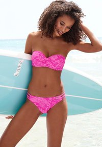 Bikini floreale rosa con top a bandeau e pantaloni con lacci laterali. Tessuto morbido, ambientazione da spiaggia, tavola da surf sullo sfondo.