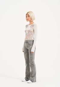 Top in rete bianco a maniche lunghe con un vivace design di tatuaggi, abbinato a jeans grigi chiari a zampa d'elefante con motivi floreali intricati e tasche laterali.
