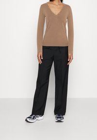 Žena v hnědém svršku s V výstřihem a detailem knoflíků, černých volných kalhotách a stříbrných a námořnických běžeckých botách, stojící na jednobarevném pozadí.