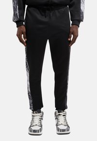 Schwarze Jogginghose mit einer taillierten Passform, versehen mit einem Seitenpanel mit einem grauen abstrakten Muster. Begleitet von gemusterten Sneakers und einem Kordelzug am Bund.