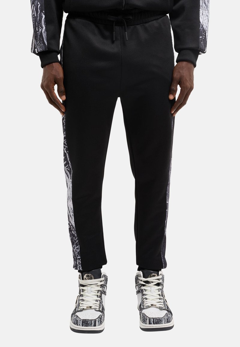 Schwarze Jogginghose mit einer taillierten Passform, versehen mit einem Seitenpanel mit einem grauen abstrakten Muster. Begleitet von gemusterten Sneakers und einem Kordelzug am Bund.