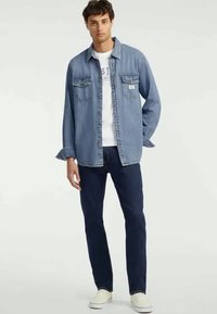 Cămașă din denim cu nasturi, de culoare albastru deschis, cu guler, două buzunare pe piept și mâneci lungi, purtată peste un tricou alb și blugi negri.