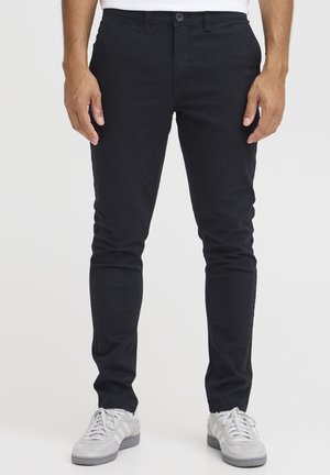 Mann trägt eng anliegende schwarze Hosen und weiße Sneaker und steht vor einem schlichten hellen Hintergrund.