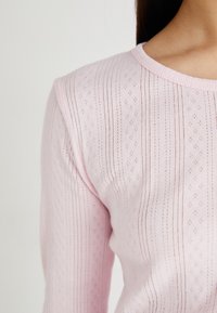Pull en tricot rose avec une texture côtelée, présentant des rayures verticales et des perforations en forme de diamant sur le tissu. Encolure ronde.