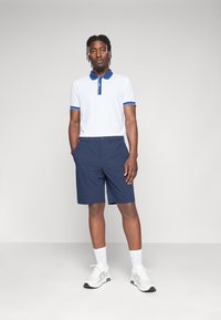 Polo shirt blanc avec col et accents bleus, associé à un short marine. Porté avec des baskets blanches et des chaussettes blanches. Texture de tissu lisse.