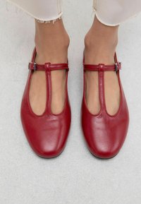 Bailarinas rojas con tira en T y acabado suave, situadas sobre una superficie gris claro, que muestran un diseño estiloso y minimalista.