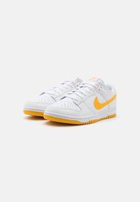 Białe skórzane sneakersy z żółtym logo Swoosh, posiadające perforowane palce, wyściełany język i gumową podeszwę z pomarańczowymi akcentami.