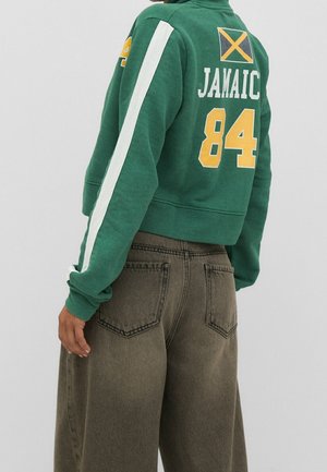 Persoon in een groene cropped sweatshirt met witte streep, gele "84," Jamaicaanse vlag en bruine wijde jeans, iets wegdraaiend.