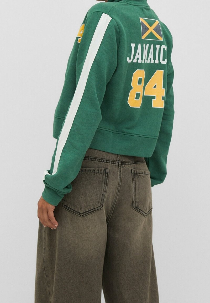 Persona che indossa un felpa verde corta con striscia bianca, il numero "84" giallo, la bandiera giamaicana e jeans a gamba larga marroni, girata leggermente di lato.