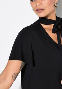 Vestito nero con maniche corte e colletto legato. Tessuto morbido, vestibilità ampia, design con scollo a V, caratterizzato da un tatuaggio sul braccio della modella.