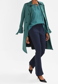 Türkiser Trenchcoat mit schwarzen Knöpfen, getragen über einer türkisen Bluse mit weißen Punkten, kombiniert mit dunkelblauen Hosen und schwarzen High Heels.