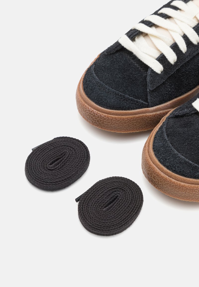 Zapatillas de ante negro con suelas de goma y cordones blancos, al lado de dos cordones negros bien enrollados sobre un fondo blanco.