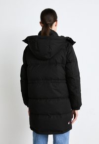 Svart pufferjakke med hette, med en quiltet design, innsvingede ermer og et logopatch ved nederkanten. Lang lengde og glatt tekstur.
