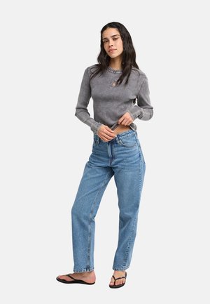 Vrouw met lang donker haar draagt een grijs shirt met lange mouwen, blauwe jeans en zwarte sandalen, staand met één hand op haar tailleband.