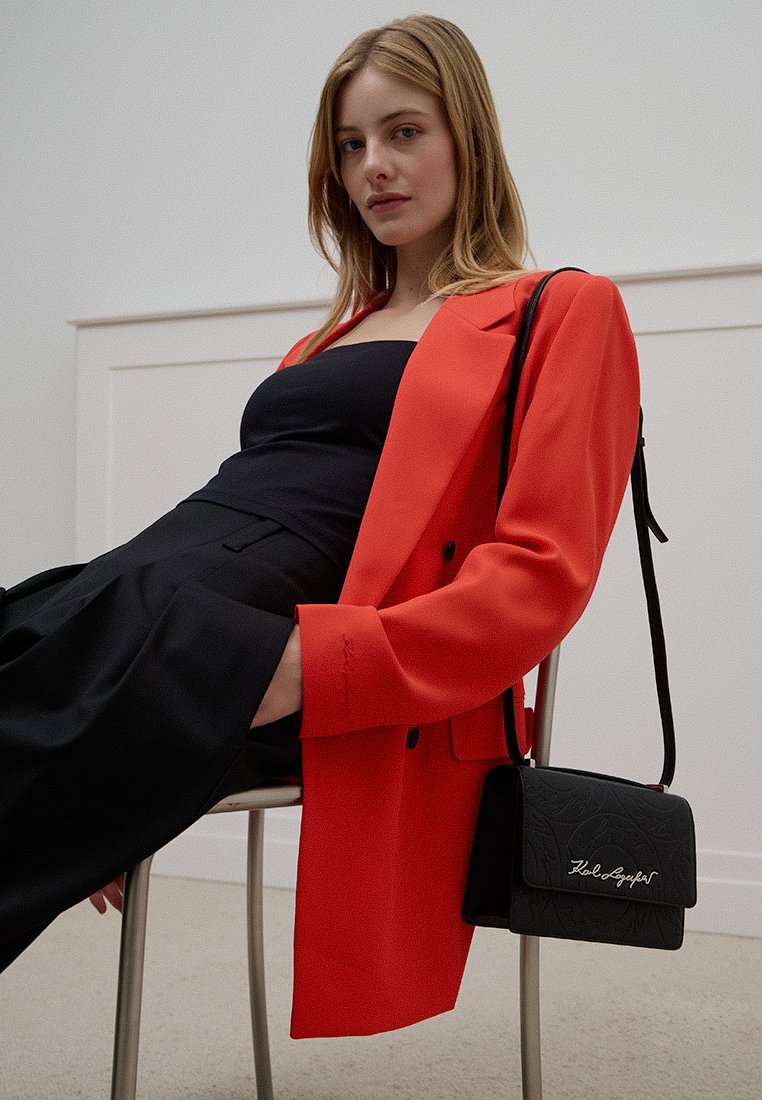 Giovane donna seduta su una sedia, indossando un blazer rosso e un outfit nero, con una borsa nera appesa sulla spalla, su uno sfondo bianco semplice.