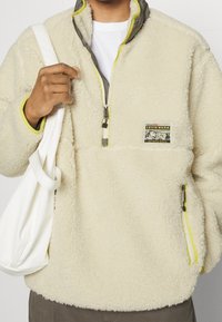 Sudadera de forro polar crema con cuello alto, cremallera frontal y ribetes amarillos en contraste. Cuenta con un bolsillo en el pecho con marca y cremalleras en los laterales.