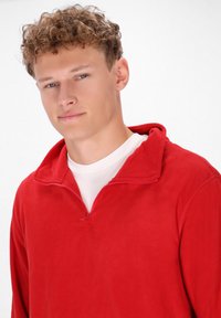 Rød fleecepullover med halv-zip krave, der har en blød tekstur og en kontrasterende hvid inderside. Maskin-sydede kanter og afslappet pasform.