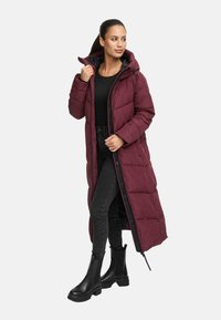 Vrouw in een lange bordeauxrode pufferjas met capuchon, zwart topje, zwarte skinny jeans en zwarte chunky boots, staand met één hand op de kraag van de jas.