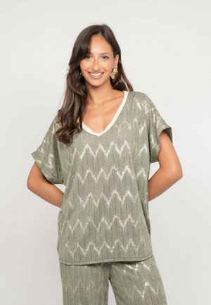 Femme souriante aux longs cheveux foncés, portant un haut à col en V à motif zigzag brillant vert olive et un pantalon assorti, debout devant un fond uni.