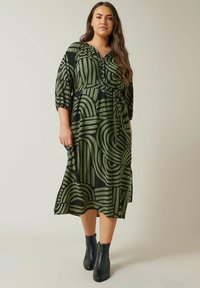 Grün-schwarz gemustertes Midi-Kleid mit Dreiviertelärmel, Taillenknoten und geometrischen Designs. Kombiniert mit schwarzen Knöchelstiefeln.