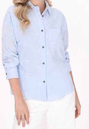 Femme portant une chemise boutonnée bleu clair brodée de fleurs avec manches retroussées et pantalon blanc sur fond blanc.