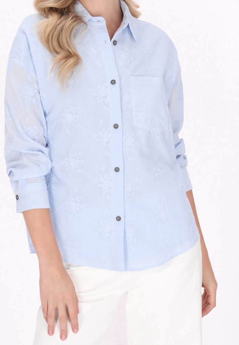 Femme portant une chemise boutonnée bleu clair brodée de fleurs avec manches retroussées et pantalon blanc sur fond blanc.