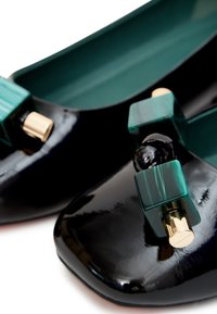 Ballerines noires en vernis avec un accent vert carré et des accessoires en métal doré. Surface lisse avec un design à bout rond.