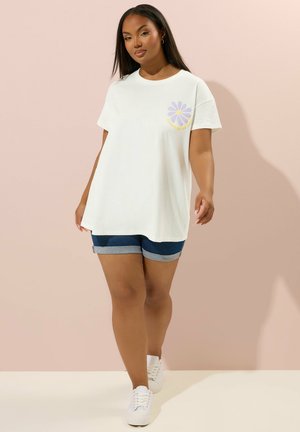 Frau trägt weißes, übergroßes T-Shirt mit lila Gänseblümchen-Motiv, dunkelblaue hochgekrempelte Jeansshorts und weiße Sneaker, geht an einer blassrosa Wand entlang.