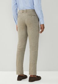 Uomo che indossa pantaloni beigi slim-fit, camicia da uomo a righe blu e mocassini scamosciati marrone scuro, in piedi contro uno sfondo chiaro e uniforme.