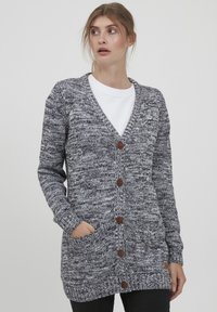 Cardigan en tricot dans un motif noir et blanc chiné, avec un décolleté en V profond, six boutons marron et deux poches avant.