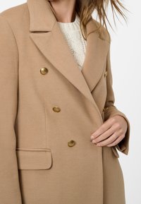 Abrigo cruzado camel en tejido texturizado, con botones dobles en tono dorado y solapa con muesca, combinado sobre un suéter de punto color crema.