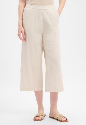 Femme portant un pantalon beige large court avec poches et des sandales beige à enfiler, debout sur fond blanc.