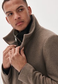 Brauner Wollmischpullover mit hohem Kragen und Reißverschluss. Verfügt über eine strukturierte Oberfläche und ein schwarzes Innenfutter, das mit den Händen am Kragen gehalten wird.