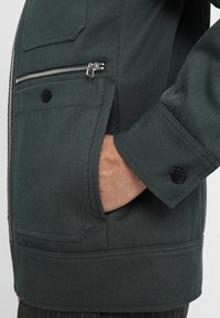 Dunkelgrüne Cordjacke mit einer Reißverschlusstasche auf der Brust, geknöpften Manschetten und einer Seitentasche. Verfügt über eine vertikale Textur und robusten Stoff.