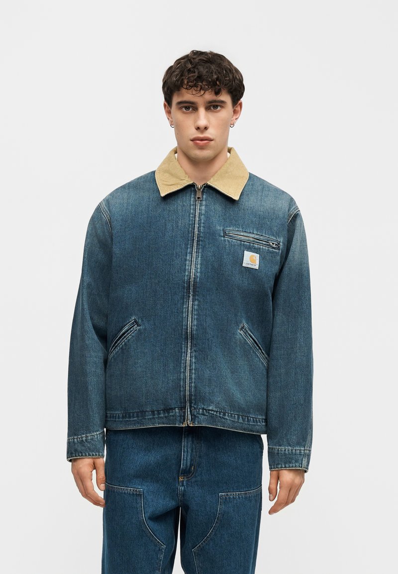 Giovane uomo con indosso una giacca di jeans blu con colletto beige e jeans di jeans blu coordinati, in piedi davanti a uno sfondo semplice.