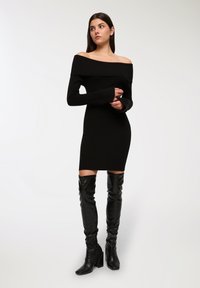 Abito nero a spalle scoperte in maglia con maniche lunghe, design aderente e stivali neri in pelle con tacco al ginocchio, caratterizzati da un aspetto texture.