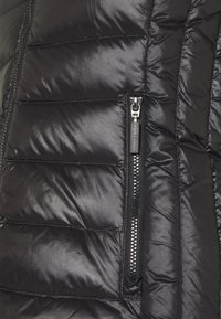 Dettaglio di giacca imbottita nera con motivo trapuntato, texture lucida e tasca con zip con ferramenta metallica ed etichetta di marca.