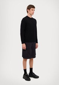 Maglione nero lavorato a maglia con motivo testurizzato, abbinato a pantaloni cargo scuri. Il modello indossa sneakers nere e calzini. Silhouette semplice e aderente.