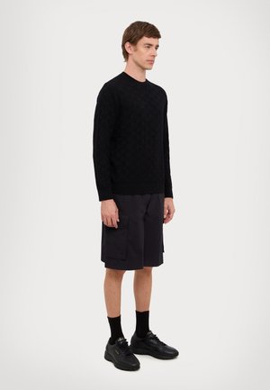 Maglione nero lavorato a maglia con motivo testurizzato, abbinato a pantaloni cargo scuri. Il modello indossa sneakers nere e calzini. Silhouette semplice e aderente.