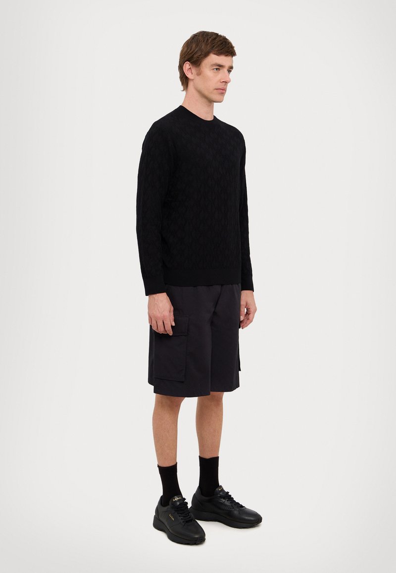 Maglione nero lavorato a maglia con motivo testurizzato, abbinato a pantaloni cargo scuri. Il modello indossa sneakers nere e calzini. Silhouette semplice e aderente.